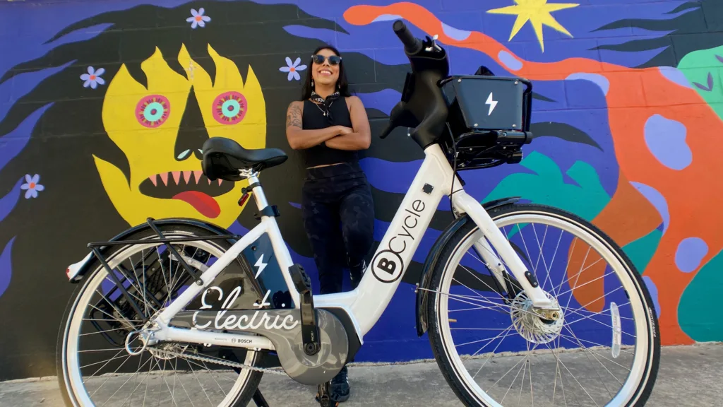 Latina woman leaning against a wall painted with a mural and her bike share bicycle is standing in front of her