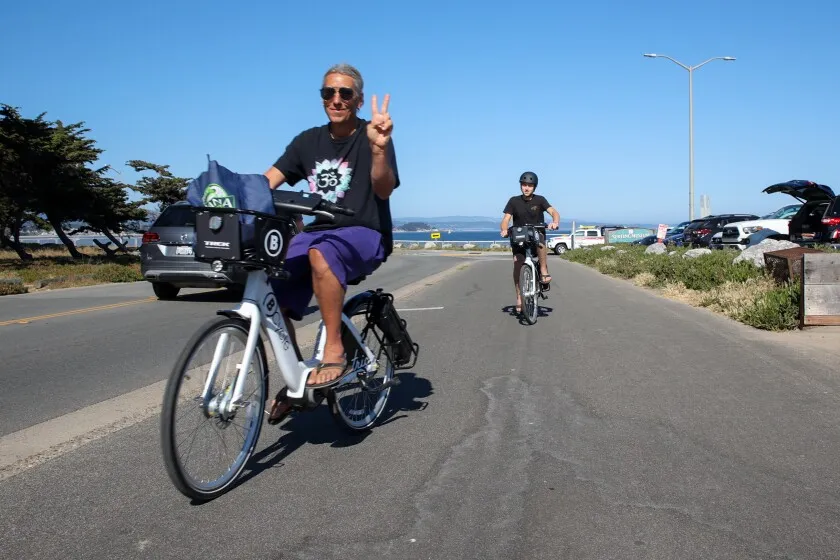 man riding a bike share bicycle on the roads of Santa Cruz giving the camera a peace sign.